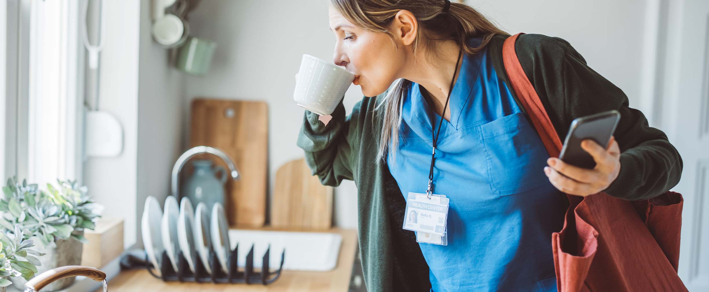 A medical professional wearing scrubs and a jacket having one last sip of coffee before leaving home