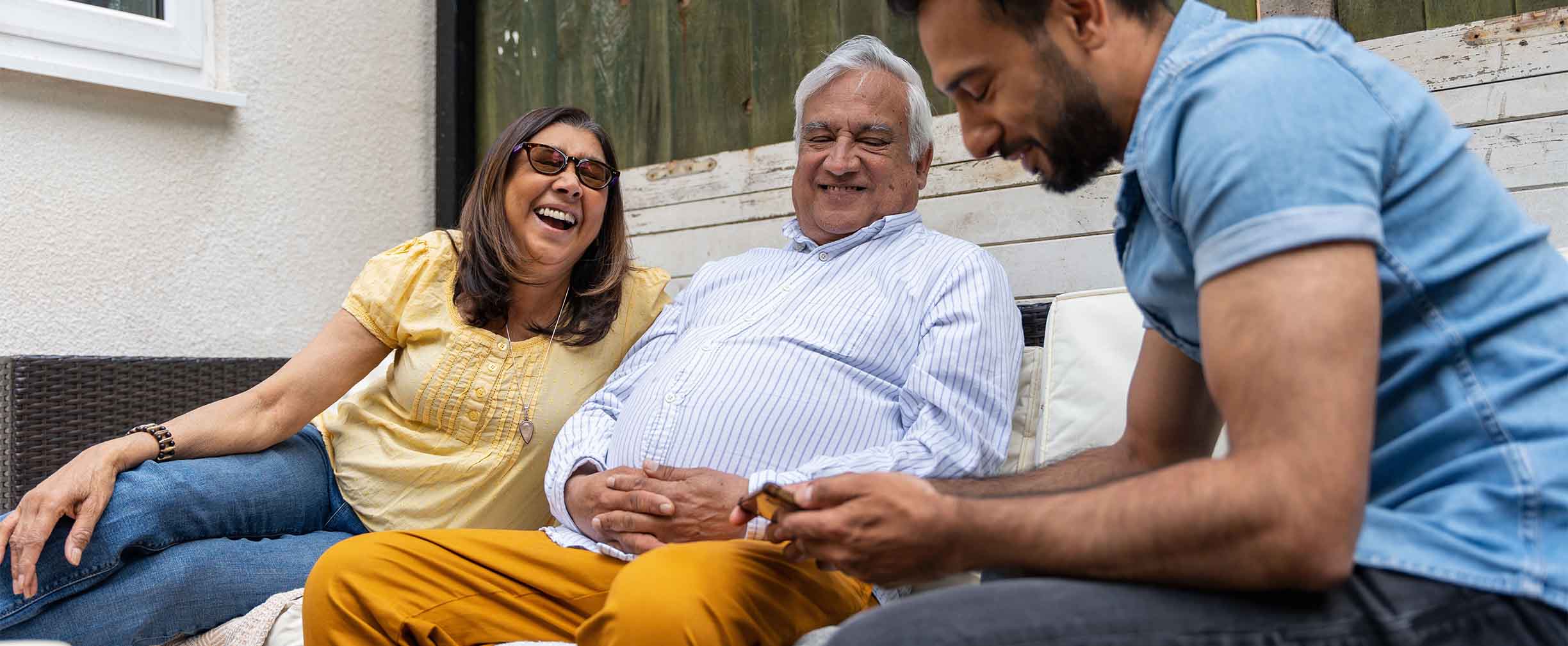 Un adulto joven riendo y sonriendo con dos familiares mayores en un patio rústico